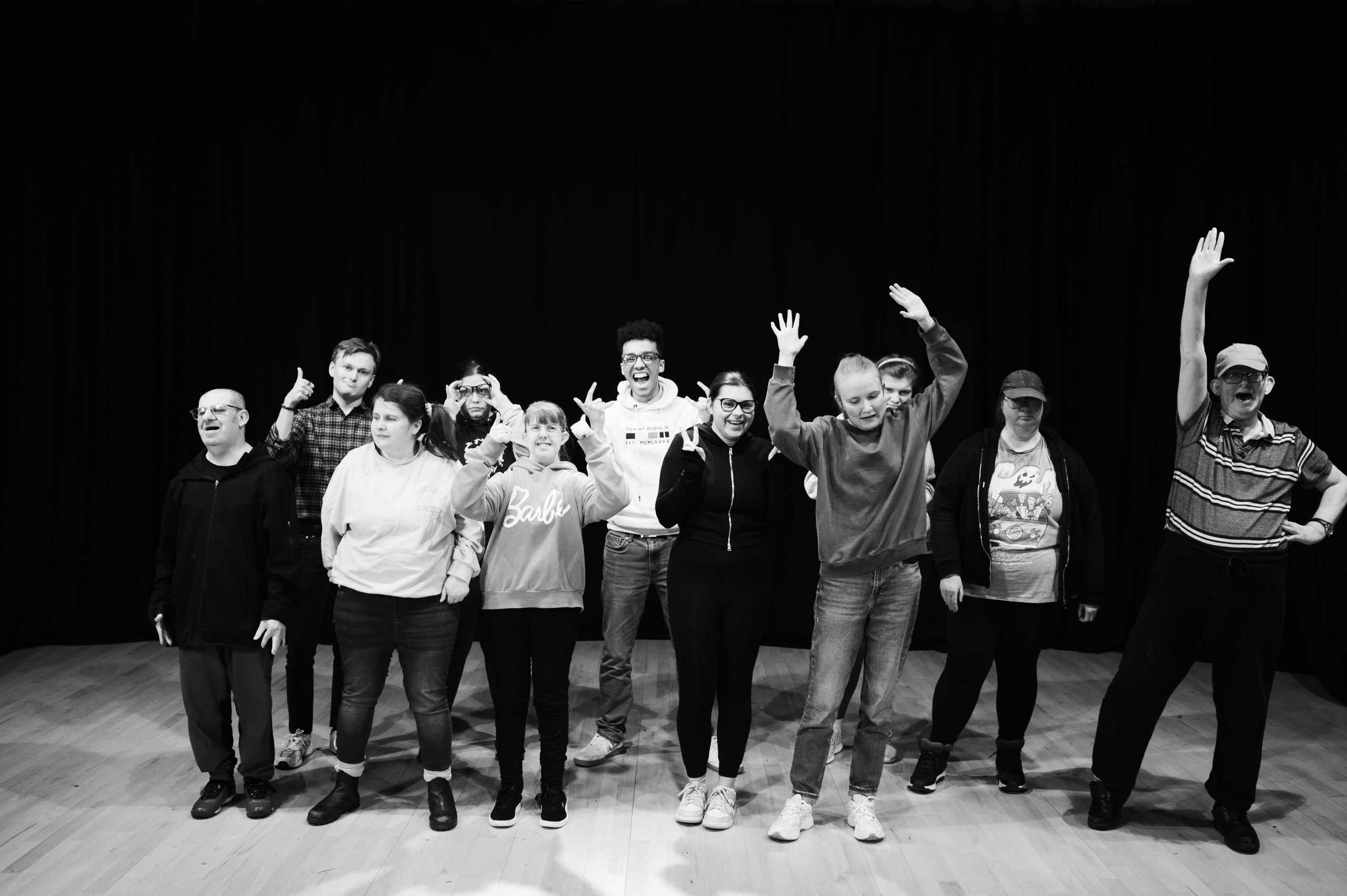 Group of diverse adults standing on stage, some smiling and raising hands, against a black backdrop in a well-lit room.